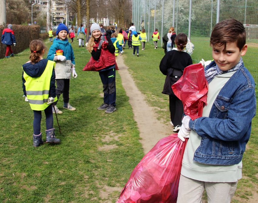 Kinder im Hintergrund mit Warnwesten gehen entlang einer Sportanlage auf der Wiese und sammeln Müll ein, im Vordergrund ein Kind mit einem großen, roten gefülltem Müllsack