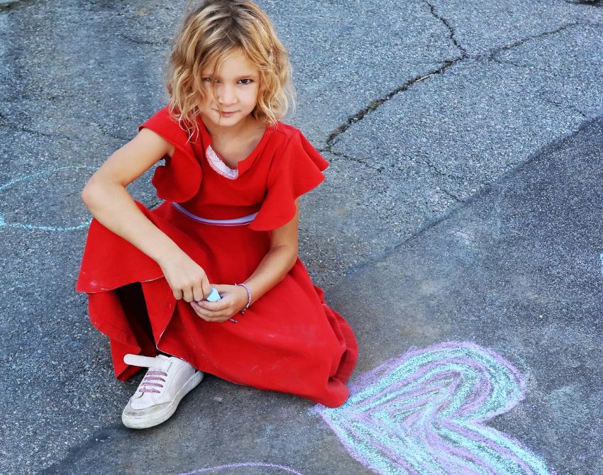Kind mit rotem Kleid sitzt auf Asphalt mit Malkreide in der Hand, neben einem mit Kreide gemaltem Herz