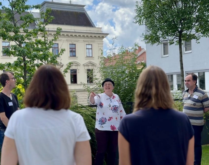 Rücken von zwei im Freien stehenden Personen, dazwischen eine Frau mit Hut sichtbar die in die Luft deutet. Dahinter ein Haus und bewölkter Himmel, Bäume am Rand