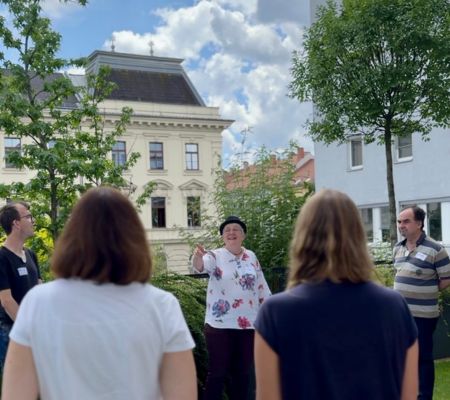 Rücken von zwei im Freien stehenden Personen, dazwischen eine Frau mit Hut sichtbar die in die Luft deutet. Dahinter ein Haus und bewölkter Himmel, Bäume am Rand