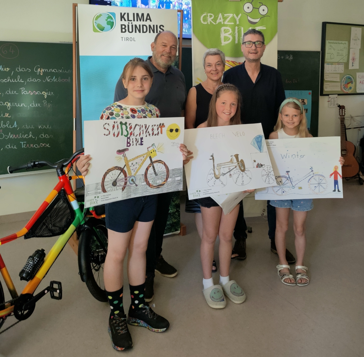 Ein buntes Fahrrad, drei Kinder stehend mit Zeichnungen von Fahrrädern und drei Erwachsene im Hintergrund in einer Schulklasse